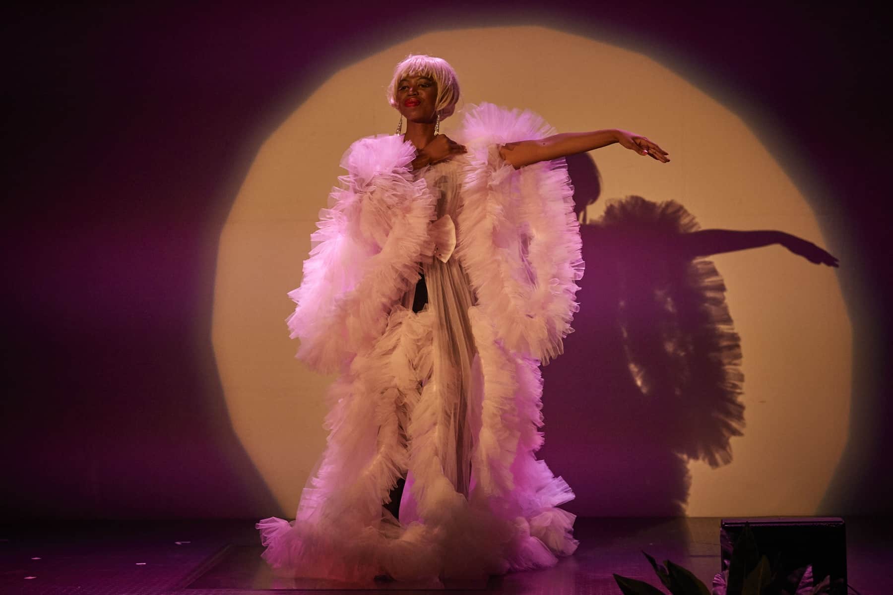 Eine Schwarze Performerin steht in einem Lichtkegel auf der Bühne. Sie trägt ein Kleid mit Rüschen und eine blonde Bubikopf-Perücke.