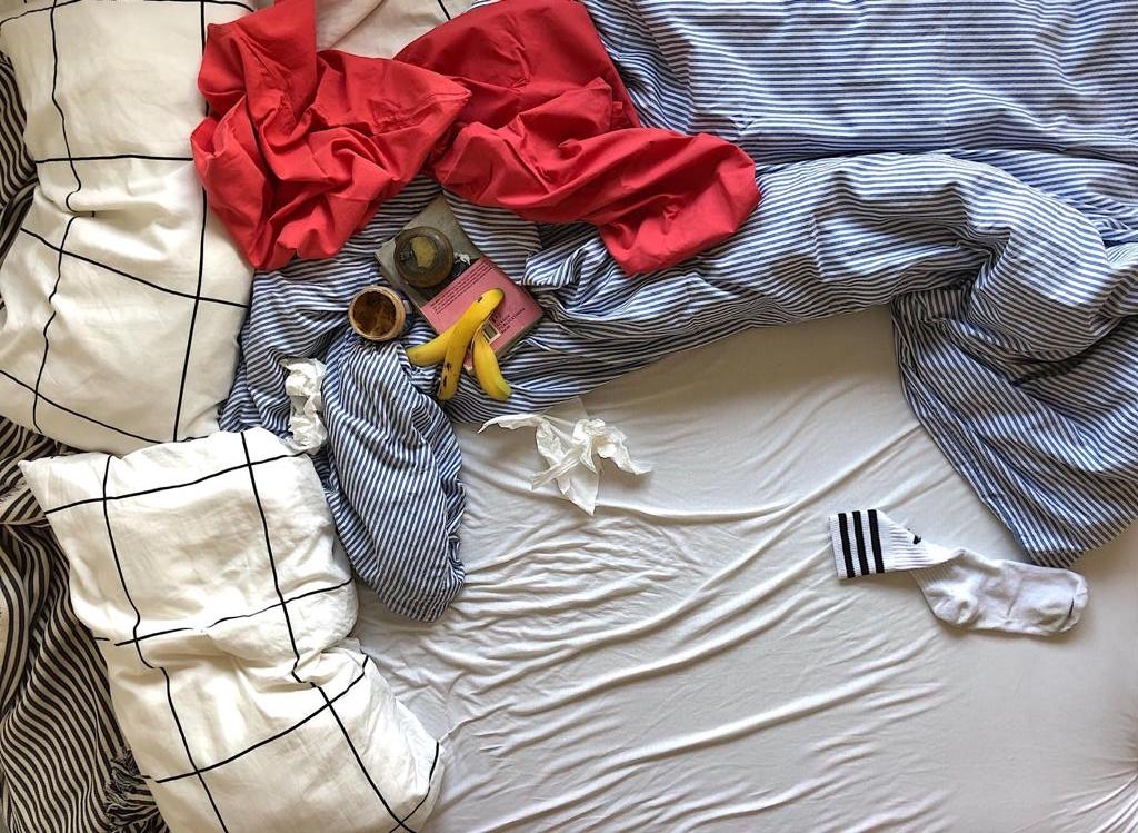 Draufsicht auf ein Bett mit unordentlicher Bettdecke. Darauf liegen ein Buch, eine Bananenschale, benutzte Taschentücher und eine einzelne Socke.