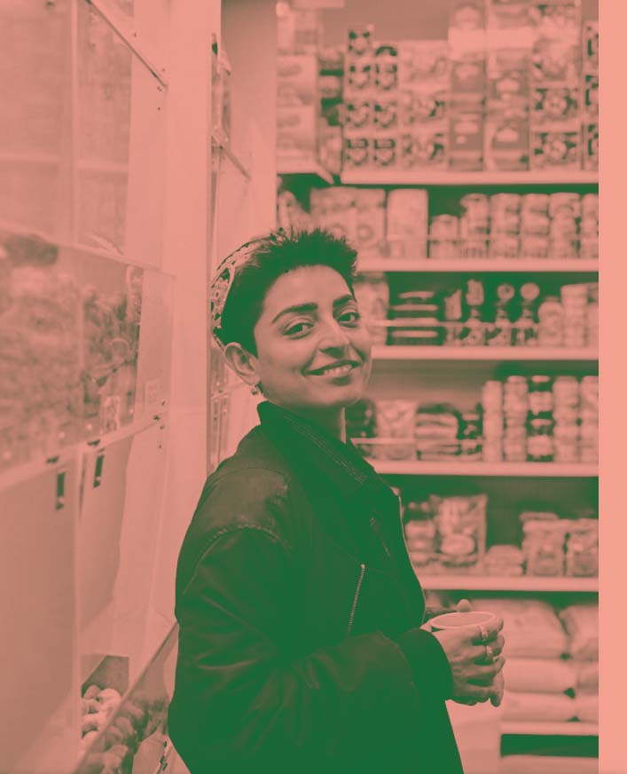Shiva Amiri steht mit einem Becher Kaffee in den Händen vor einem Lebensmittelregal und lächelt in die Kamera.