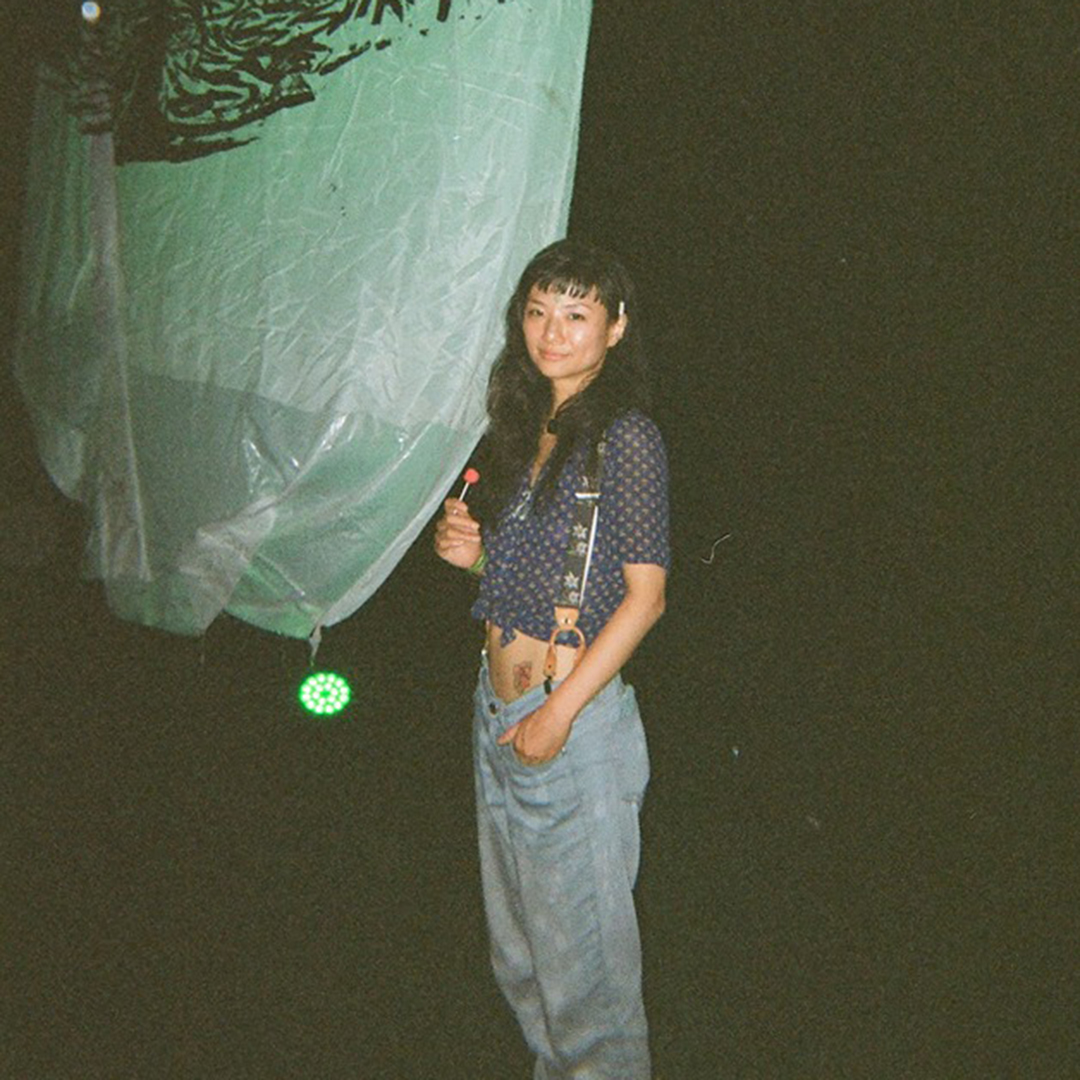Portrait von Saeko Killy im Dunkeln. Sie trägt eine Latzhose und ein bauchfreies Shirt, in der Hand hält sie einen Lolli. Hinten links ist eine Plastikplane zu sehen.
