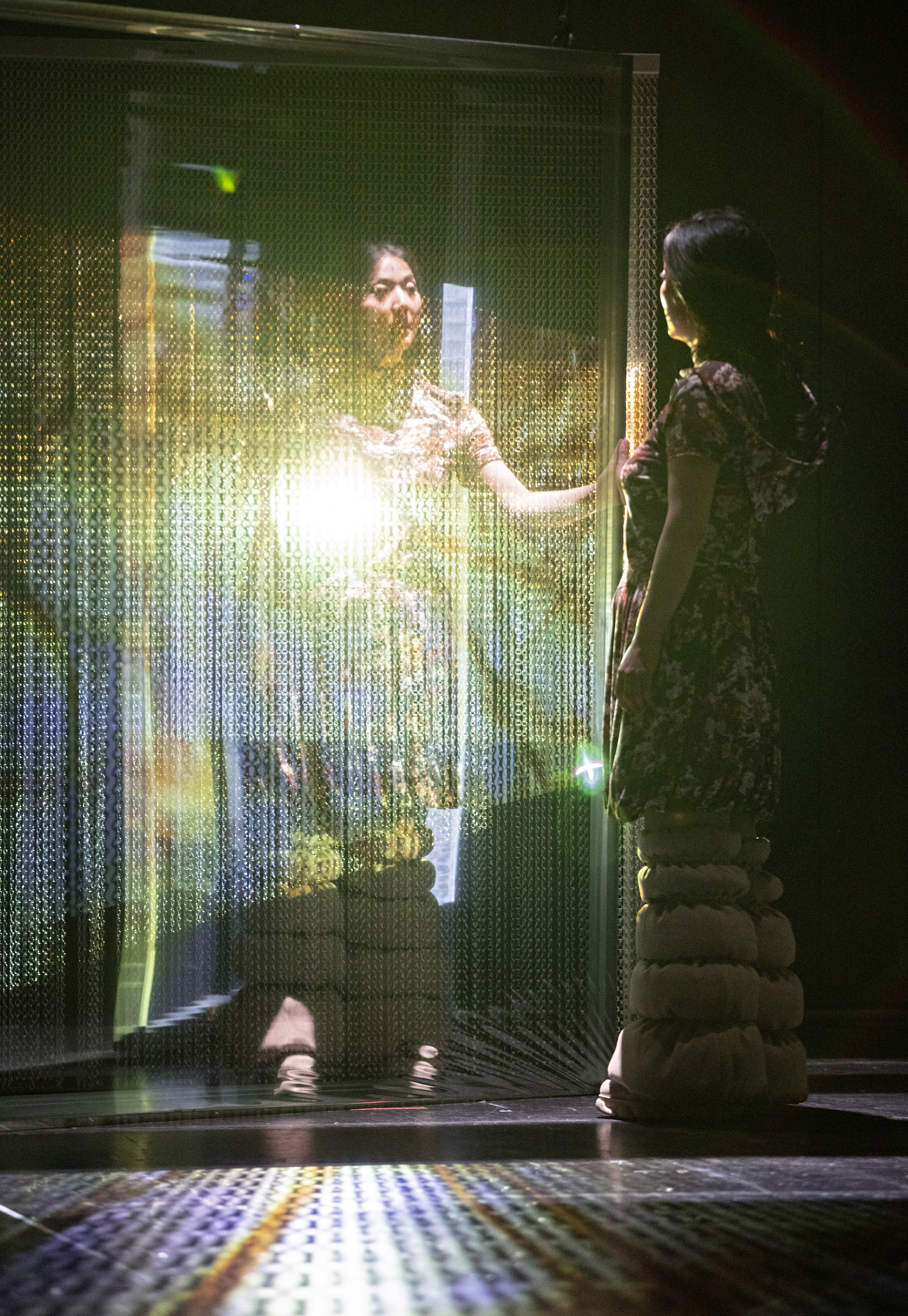 Das Foto zeigt eine Person, die im Dunkeln auf einer Bühne steht und in einen angeleuchteten Spiegel schaut. Sie steht abgewandt zur Kamera, ihr Spiegelbild der Kamera zugewandt.