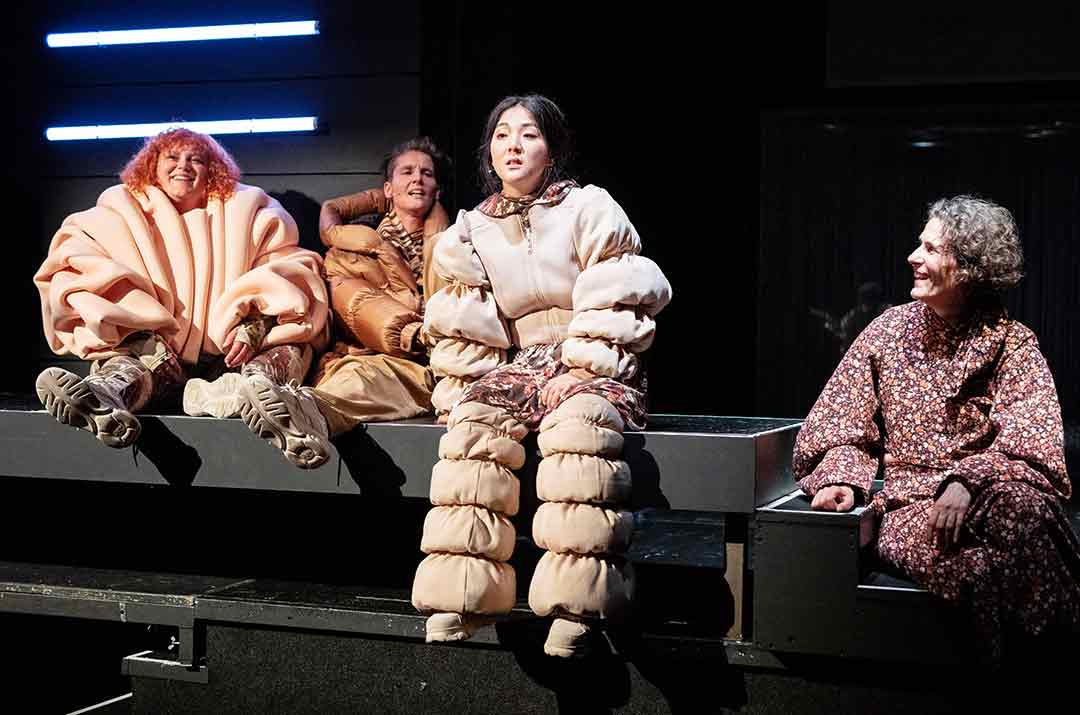 Vier Personen sitzen auf einer Bühne. Sie tragen rötlich-beige Kleidung und teilweise beige Kostüme, die kunstvoll Falten werfen.
