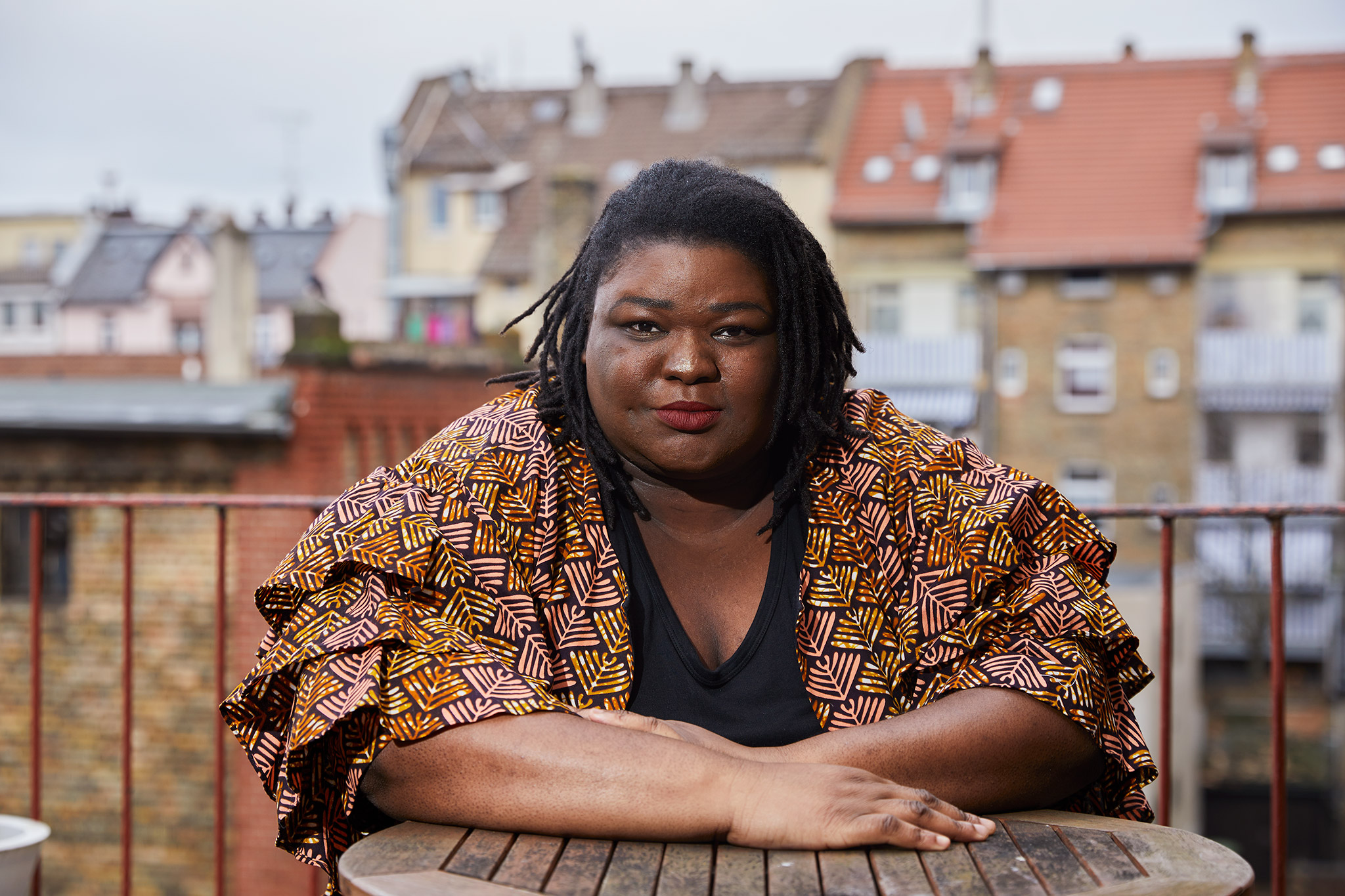 Portrait von Christelle Nkwendja-Ngnoubamdjum, einer dark-skinned Schwarzen fetten Cis-Hetero Frau auf einem Balkon vor einer Häuserfassade. Sie trägt schulterlange schwarze Rastazöpfe und eine gemusterte Jacke über einem schwarzen Shirt.