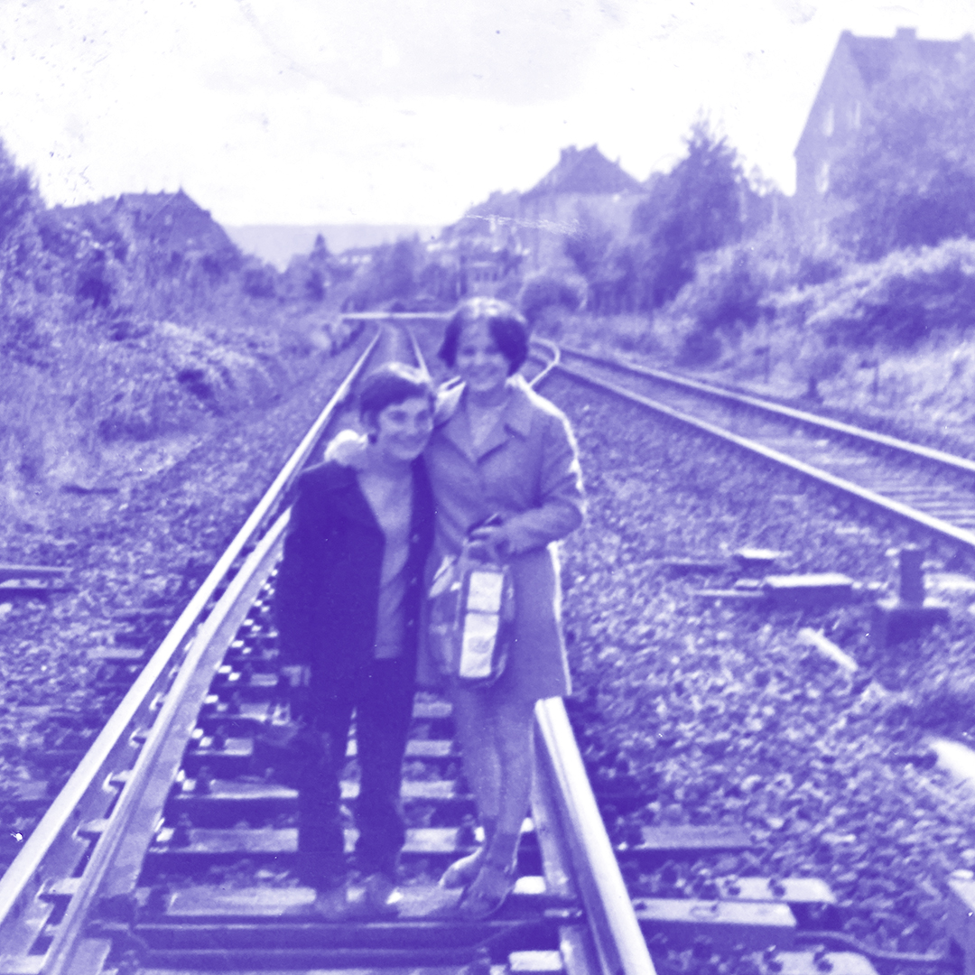 Ein in Blautönen eingefärbtes Foto, auf dem ein Kind und eine erwachsene Frau auf Bahngleisen stehen und in die Kamera lächeln. Ringum stehen Bäume und einzelne Häuser.