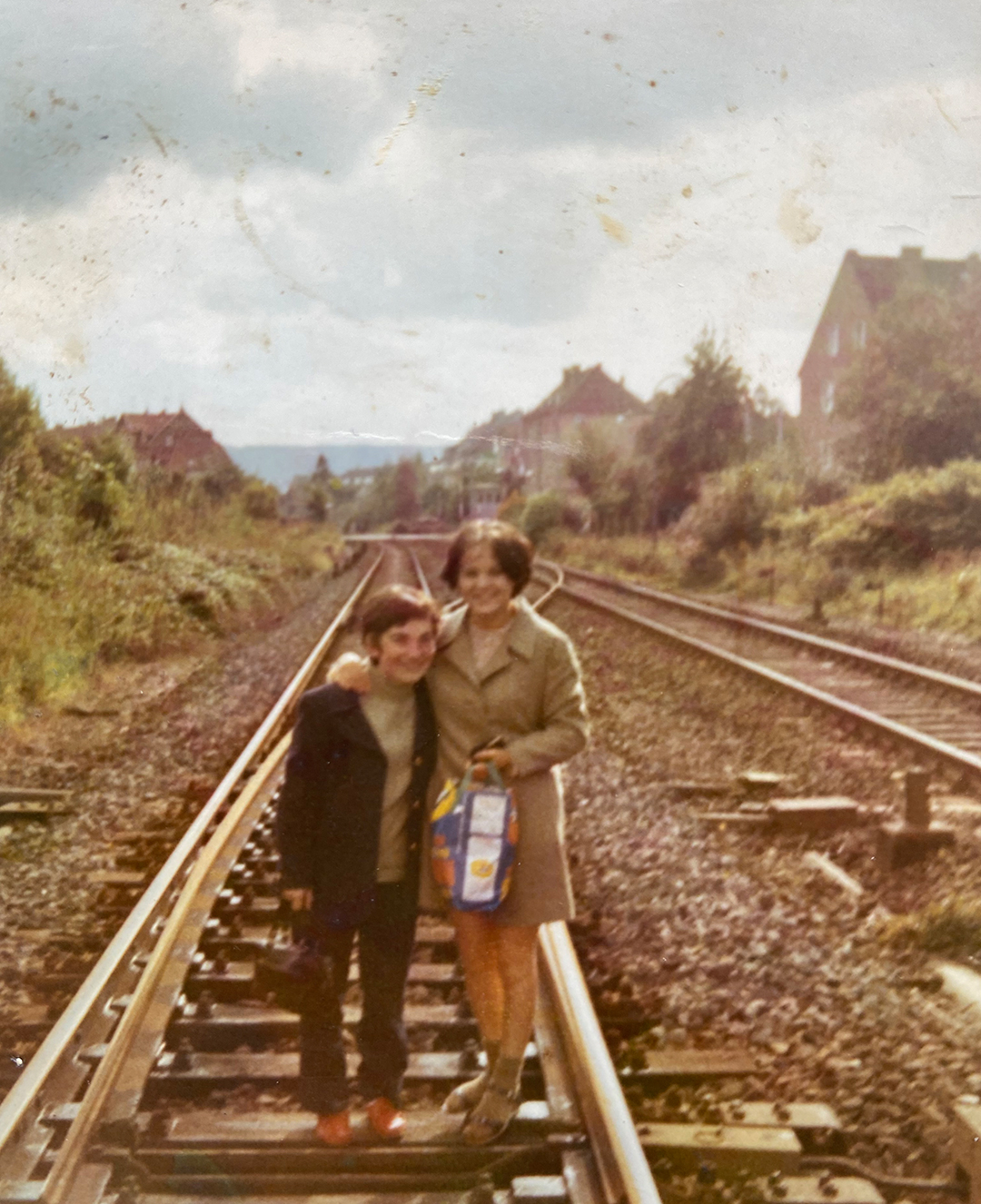 Ein altes Foto, auf dem ein Kind und eine erwachsene Frau auf Bahngleisen stehen und in die Kamera lächeln. Ringum stehen Bäume und einzelne Häuser.