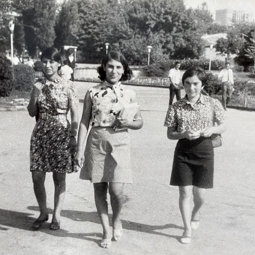 Ein schwarzweißes Foto zeigt drei Frauen im Freien, die in Richtung Kamera schauen. Alle drei tragen knielange Röcke und Blusen, die Person in der Mitte hat ein Buch im Arm.
