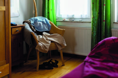 Sichtbar ist ein Teil eines Wohnraums. Mittelpunkt des Fotos ist ein Stuhl, auf dem Kleidung verteilt ist.