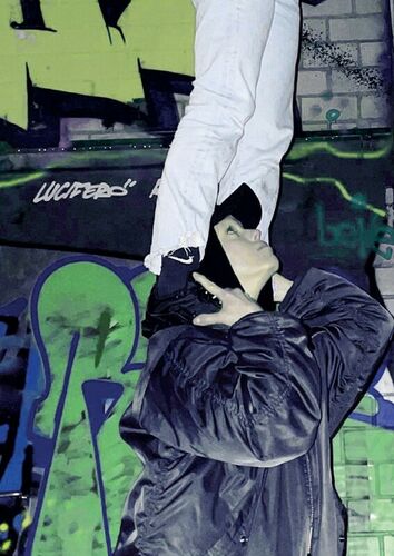 Das Foto zeigt zwei Personen vor einer mit Graffiti versehenen Wand. Von einer Person sind die Beine zu sehen, sie steht mit beiden Füßen auf den Schultern der zweiten Person, die bis zum Oberkörper auf dem Foto sichtbar ist. Die Person unten schaut nach rechts oben.